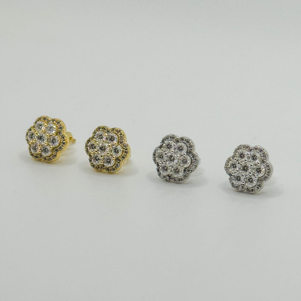 Four small flower-shaped stud earrings arranged in a row on a light gray background, showing both yellow gold and silver-tone metal options with clear pavé stones.