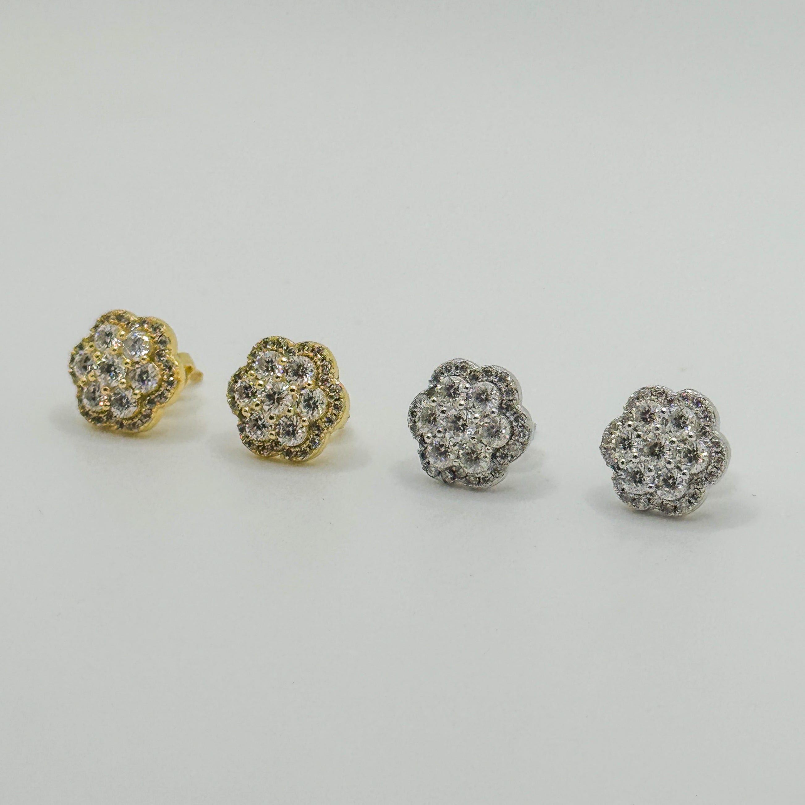 Four small flower-shaped stud earrings arranged in a row on a light gray background, showing both yellow gold and silver-tone metal options with clear pavé stones.