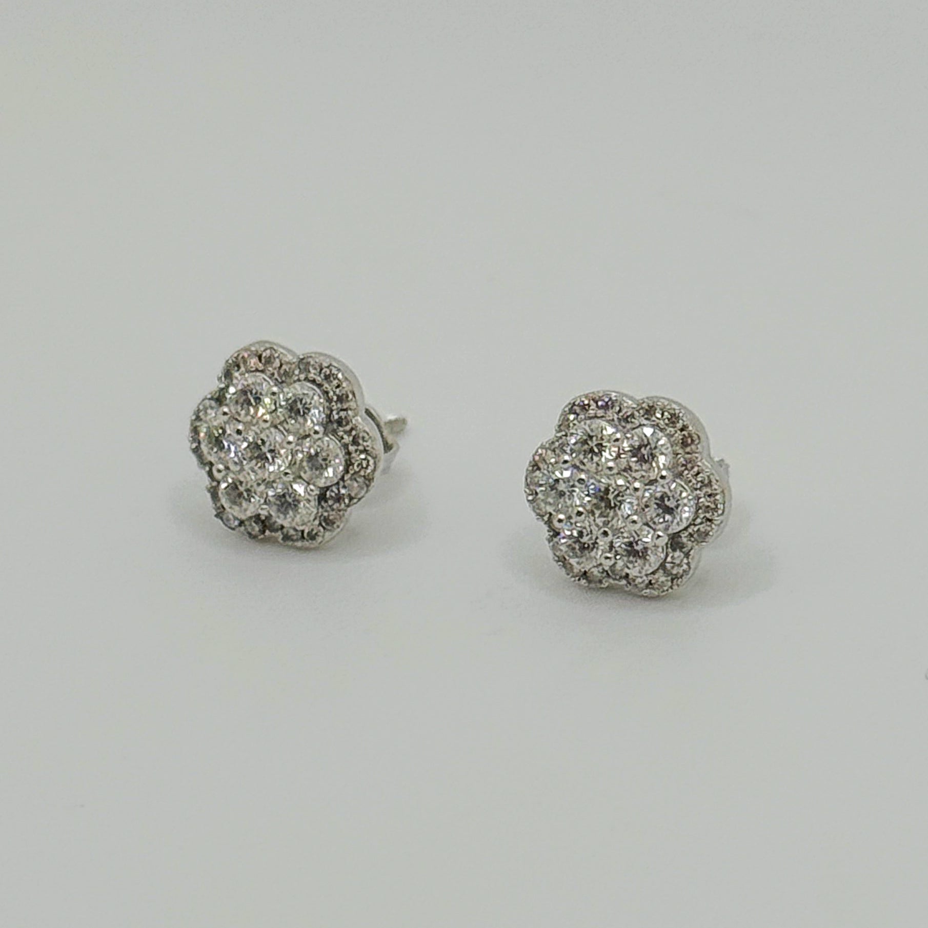 A pair of round flower-shaped stud earrings set with clear pavé gemstones in a silver-toned metal, displayed on a neutral light gray background.