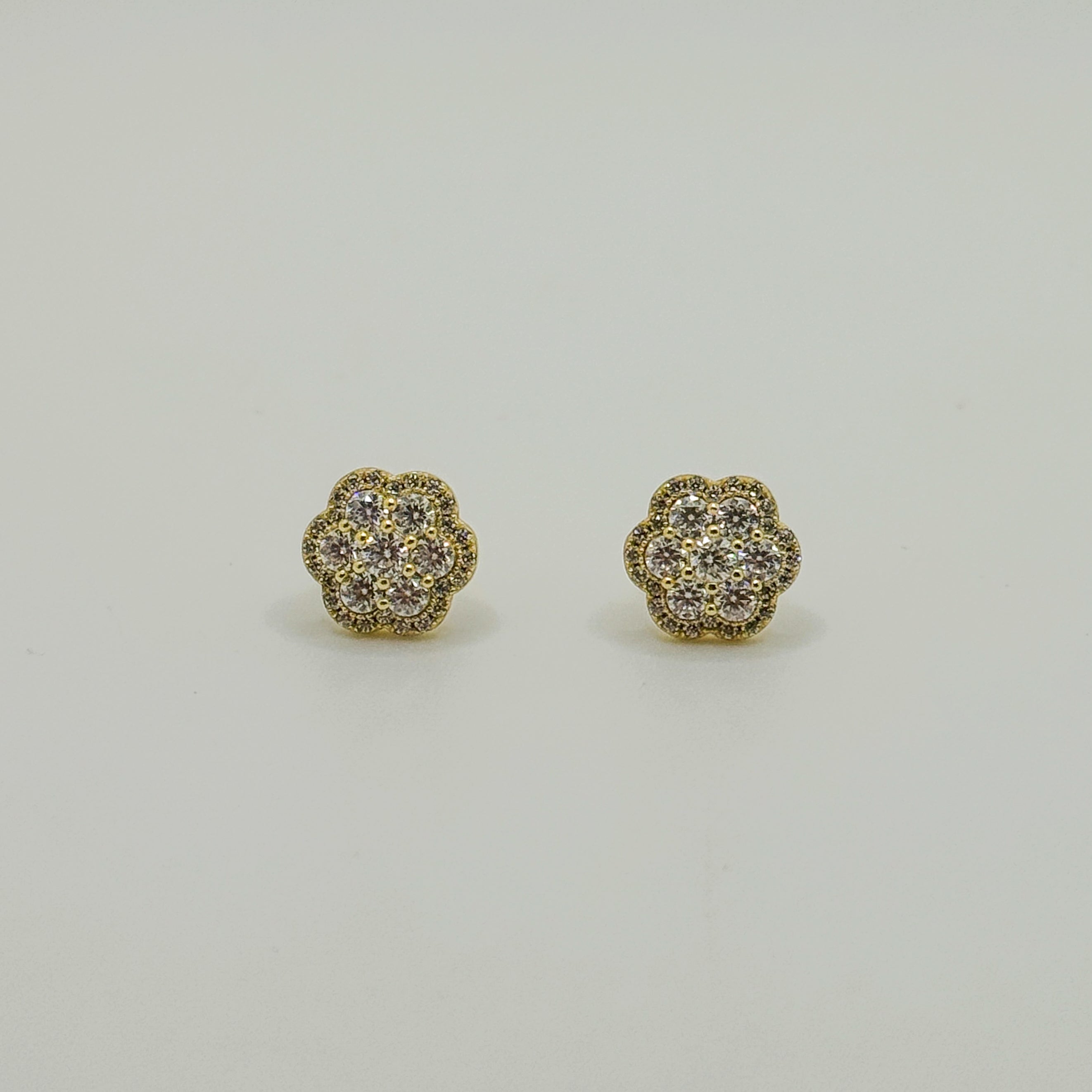A pair of gold flower-shaped stud earrings with pavé-set clear stones, displayed on a clean, light gray background.