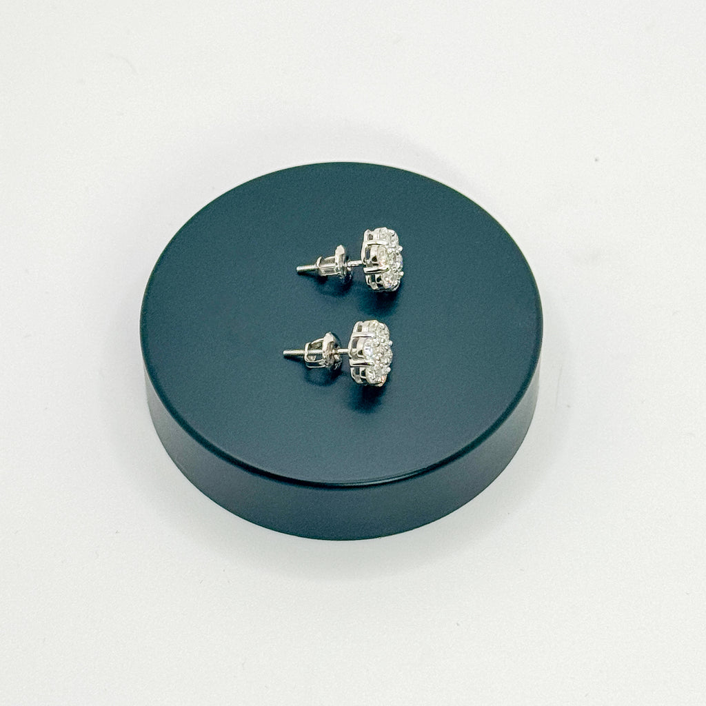 A pair of silver-toned stud earrings with clear crystal-like stones, displayed on a round dark teal stand against a plain white background.