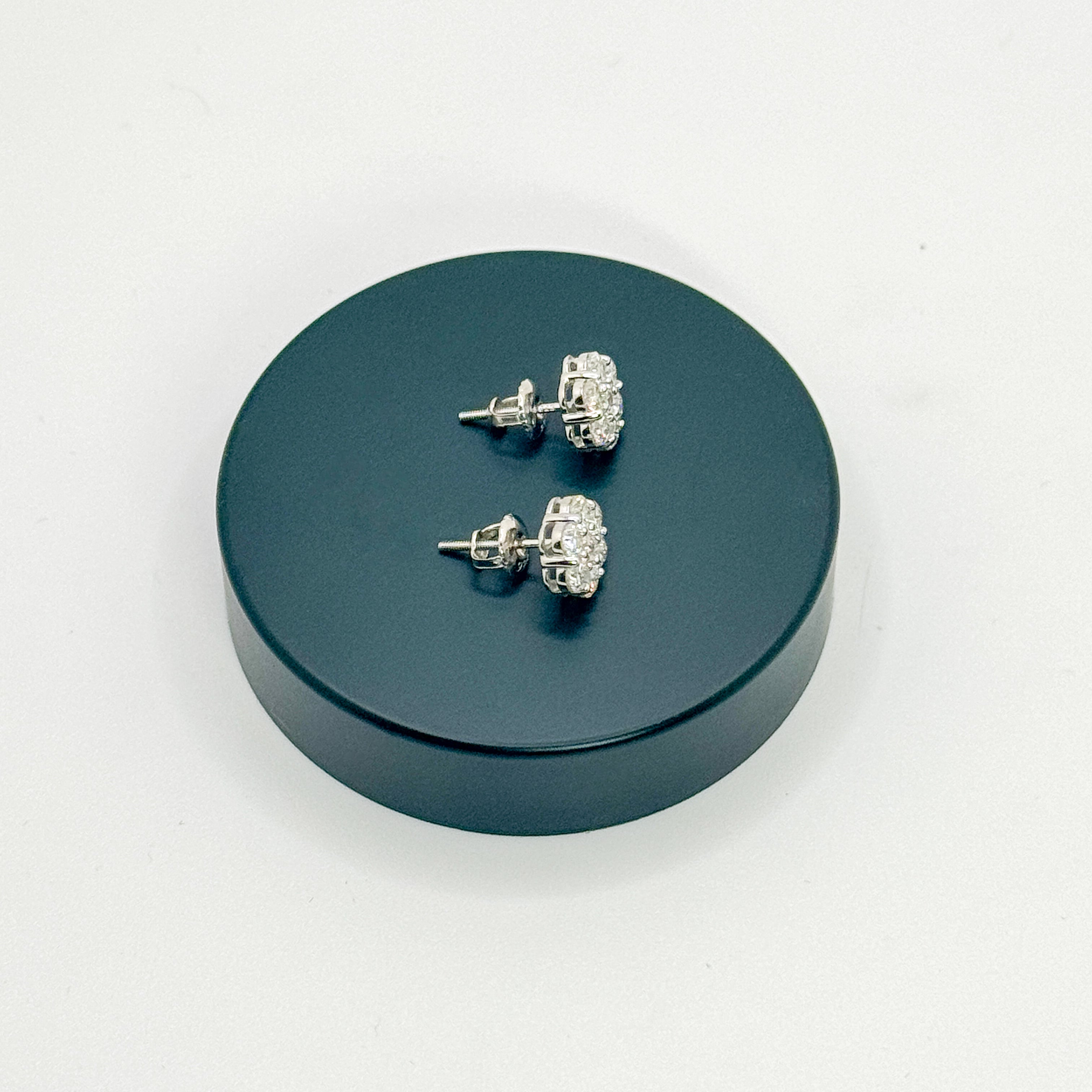 A pair of silver-toned stud earrings with clear crystal-like stones, displayed on a round dark teal stand against a plain white background.