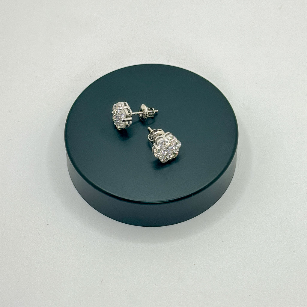 A pair of silver-toned stud earrings with clear gemstones, displayed on a round black stand against a light gray background.