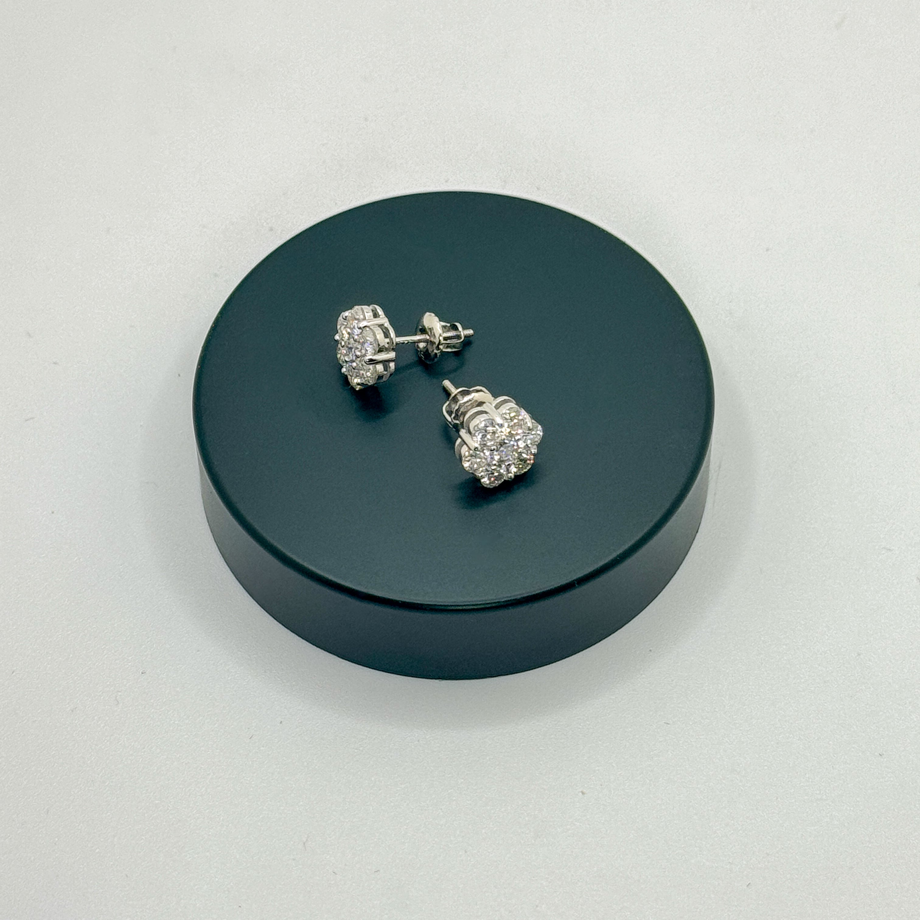 A pair of silver-toned stud earrings with clear gemstones, displayed on a round black stand against a light gray background.