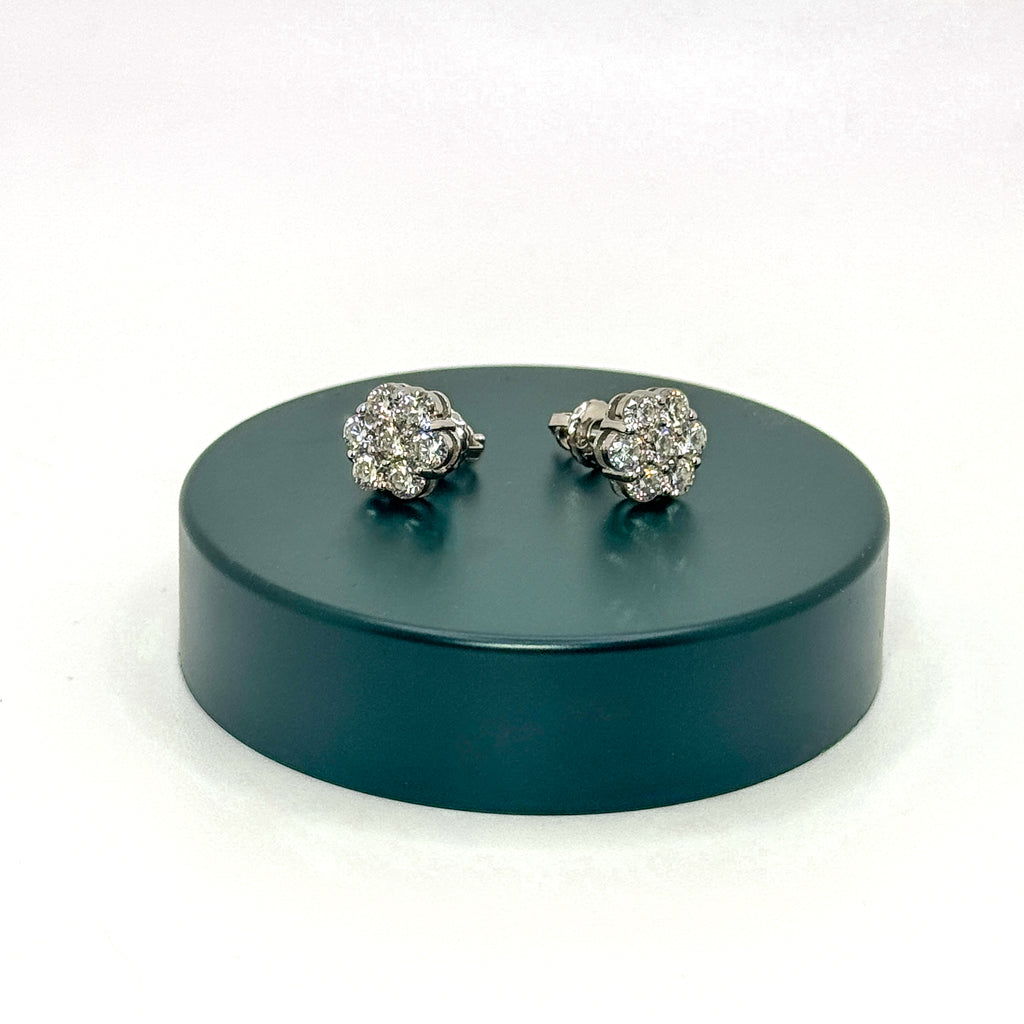 A pair of silver-toned pavé diamond stud earrings displayed on a round dark green pedestal against a white background.