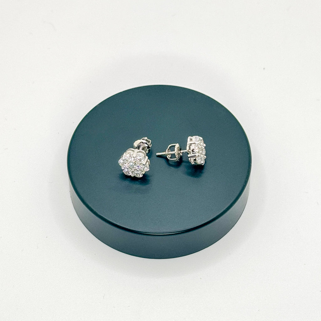 A pair of silver-toned stud earrings with clear pavé-style stones, displayed on a round dark teal stand against a plain white background.