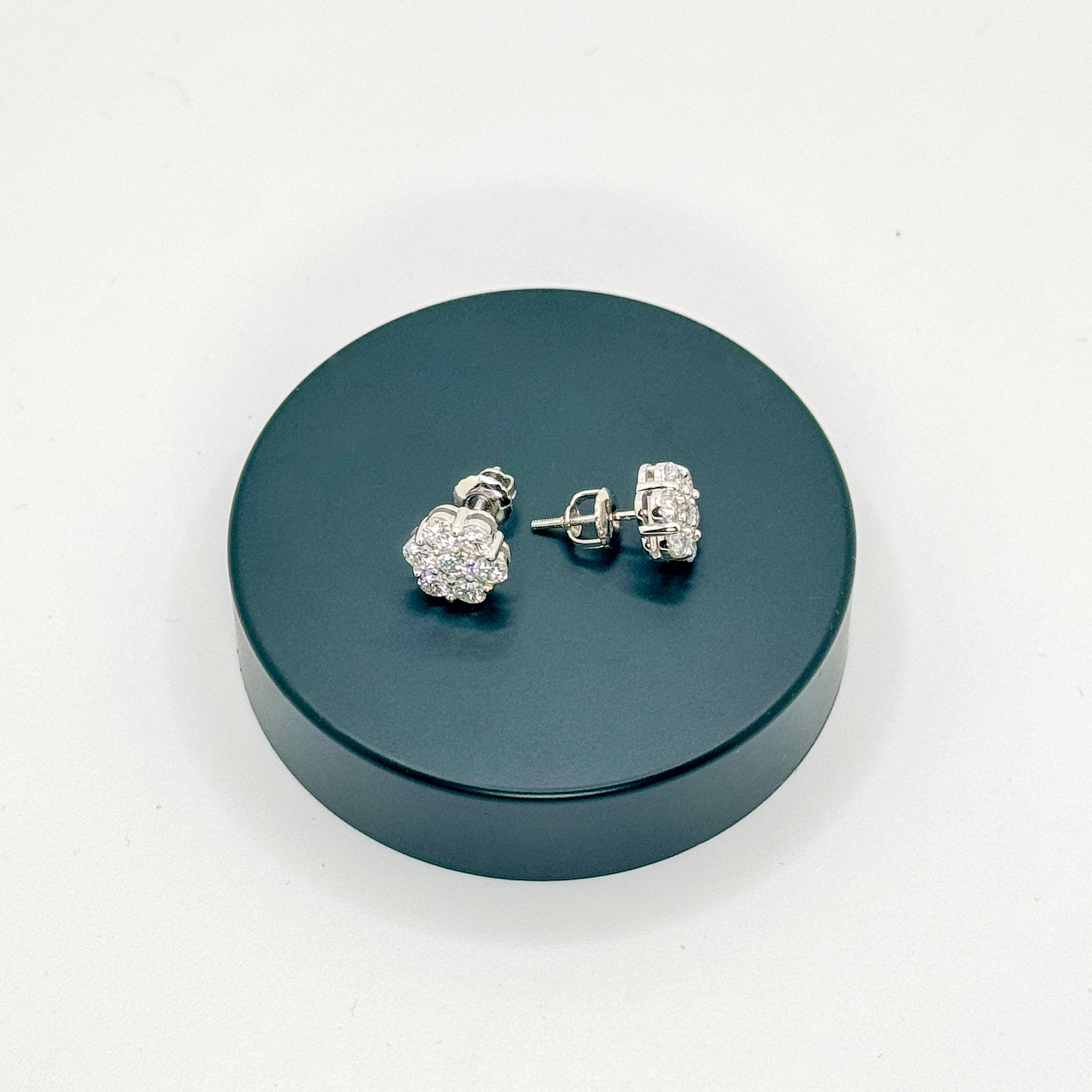 A pair of silver-toned stud earrings with clear pavé-style stones, displayed on a round dark teal stand against a plain white background.