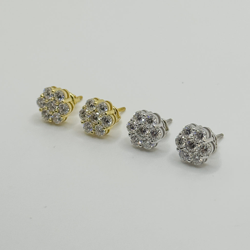 Four small flower-shaped stud earrings arranged diagonally on a light gray background, including two gold-tone and two silver-tone versions with clear gemstones.