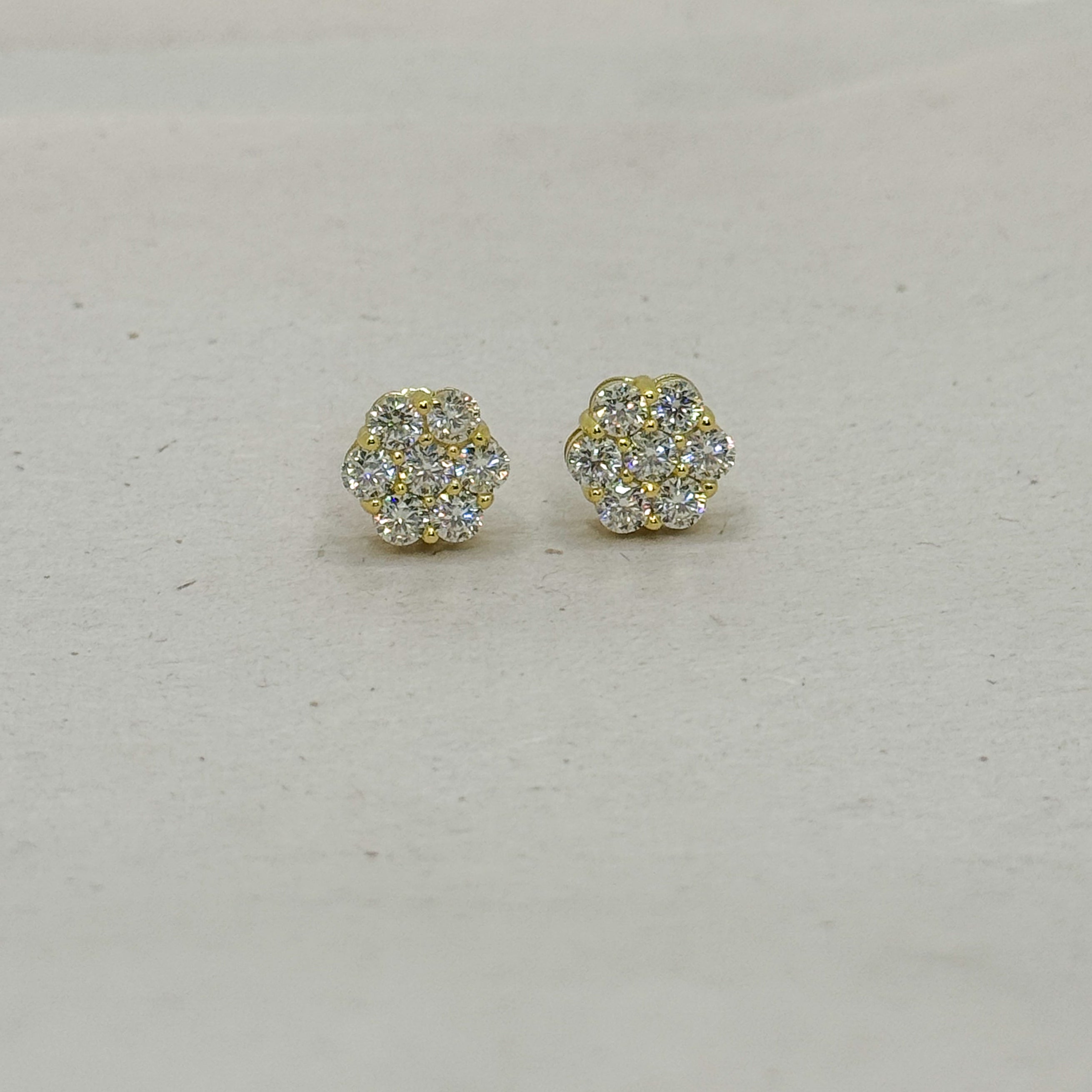 A pair of small gold stud earrings with clear pavé gemstones, arranged on a light beige textured surface.