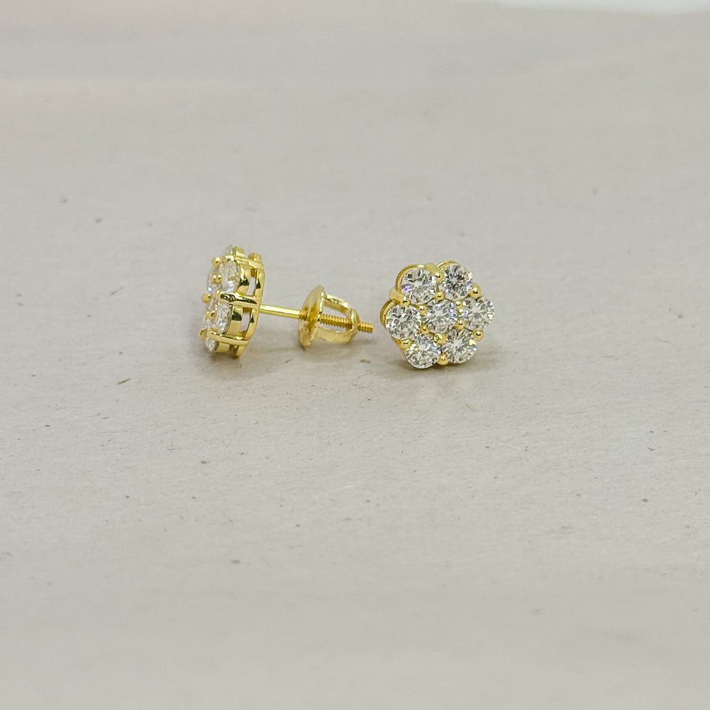 A pair of gold stud earrings, one showing the back post and the other showing the front flower-shaped cluster set with clear stones, on a light gray textured background.