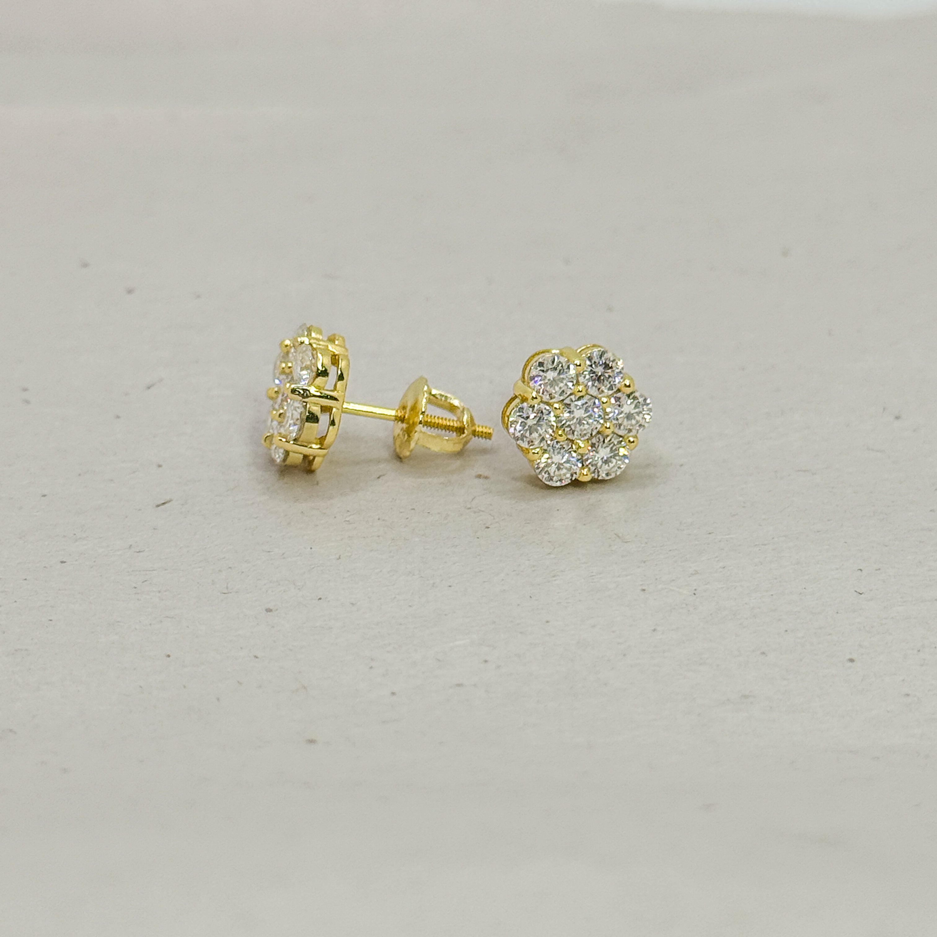 A pair of gold stud earrings, one showing the back post and the other showing the front flower-shaped cluster set with clear stones, on a light gray textured background.