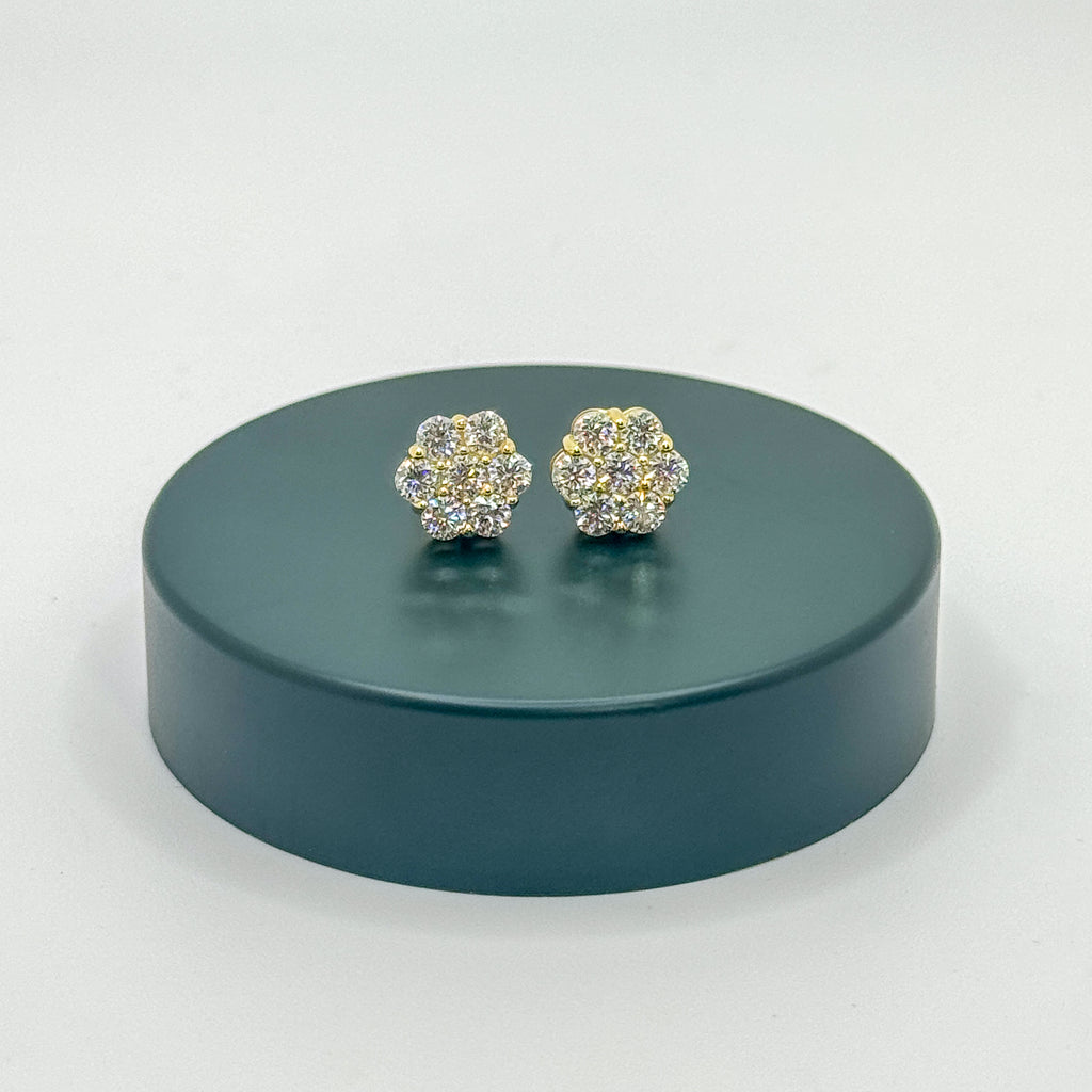 A pair of gold-toned stud earrings covered in clear crystals, displayed on a round dark teal pedestal against a light gray background.