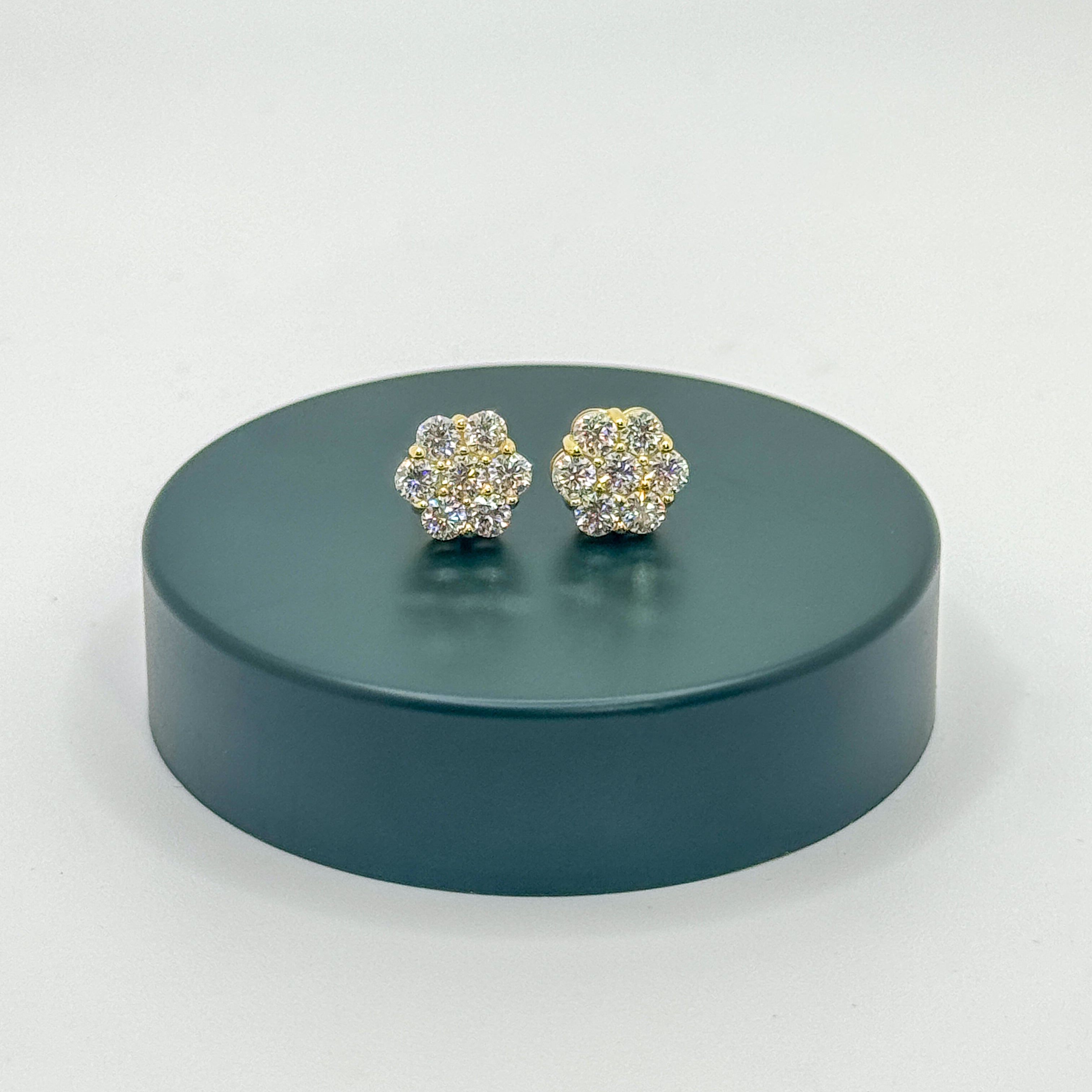 A pair of gold-toned stud earrings covered in clear crystals, displayed on a round dark teal pedestal against a light gray background.