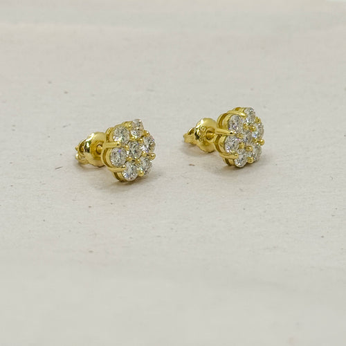 A pair of yellow gold stud earrings with round-cut clear gemstones, displayed on a light beige textured surface.