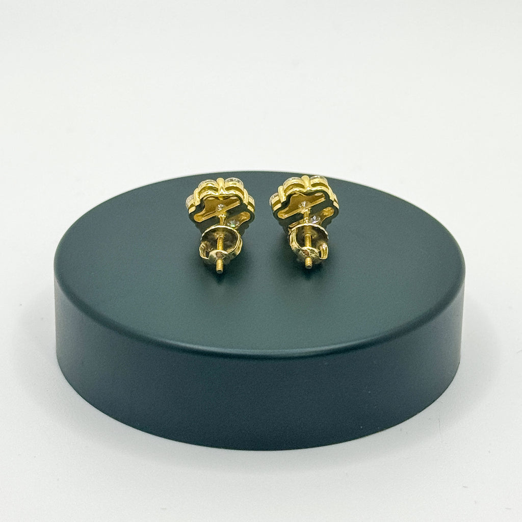 A pair of gold stud earrings with a textured, flower-like top, shown from the back on a round dark green display stand against a white background.