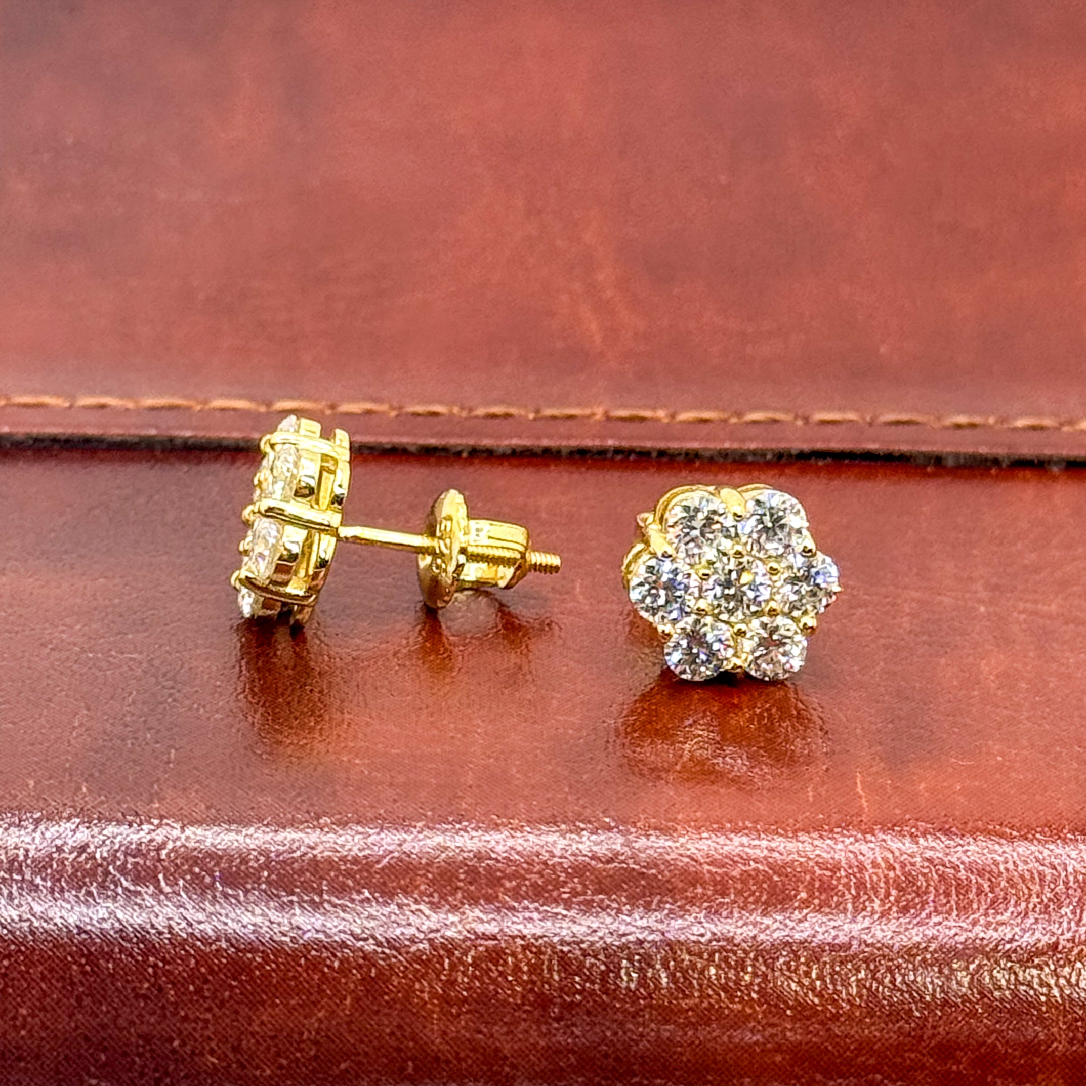 A pair of gold stud earrings displayed on a brown leather surface, with one earring shown from the side/back and the other from the front. The front earring features a cluster of clear gemstones in a flower-like shape.