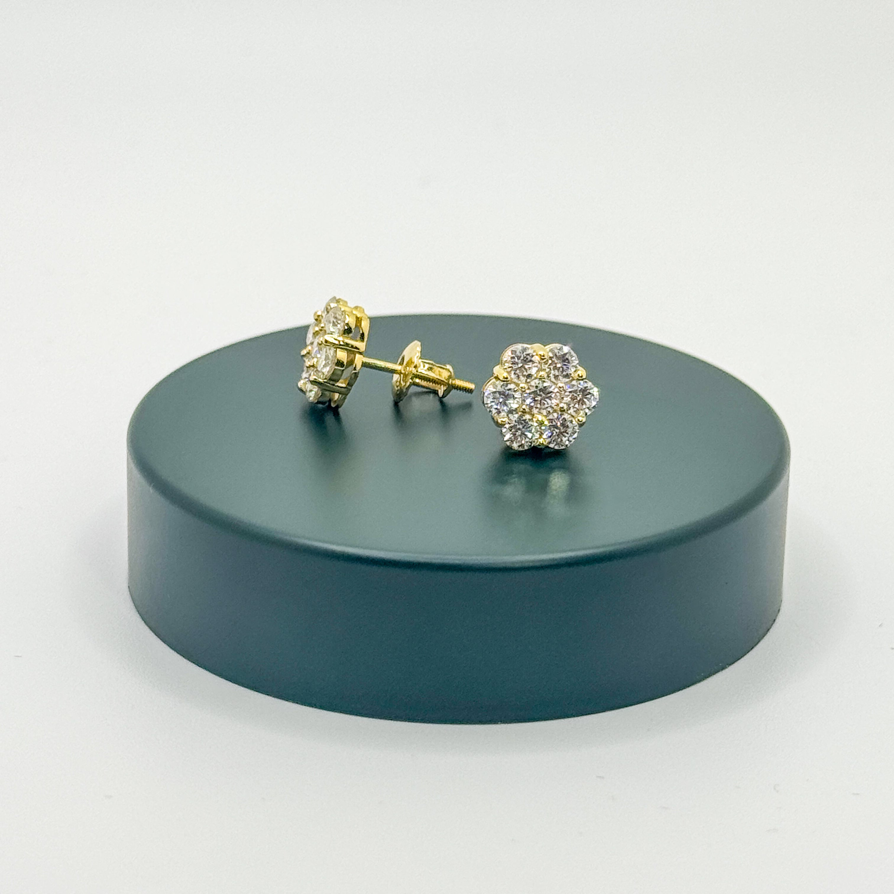 A pair of gold stud earrings with clear gemstones, displayed on a round dark green pedestal against a light gray background.