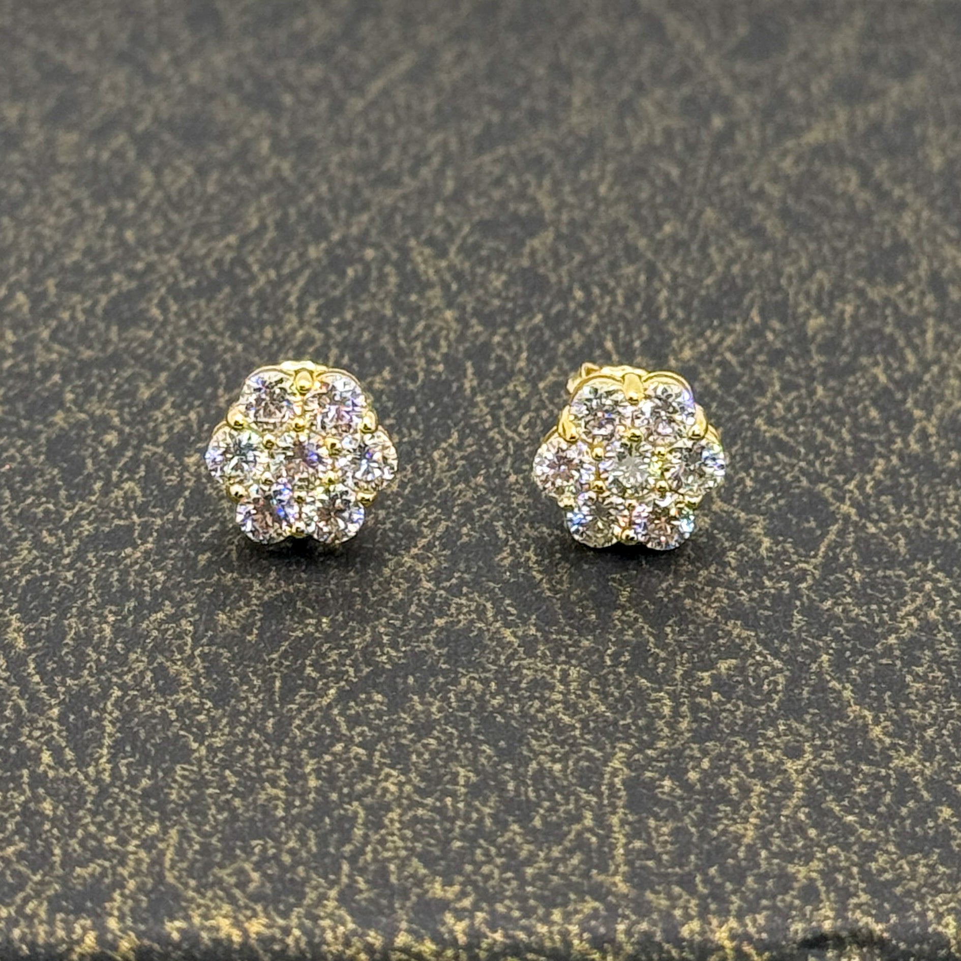 A pair of gold stud earrings with clear pavé gemstones, displayed on a textured dark gray surface.