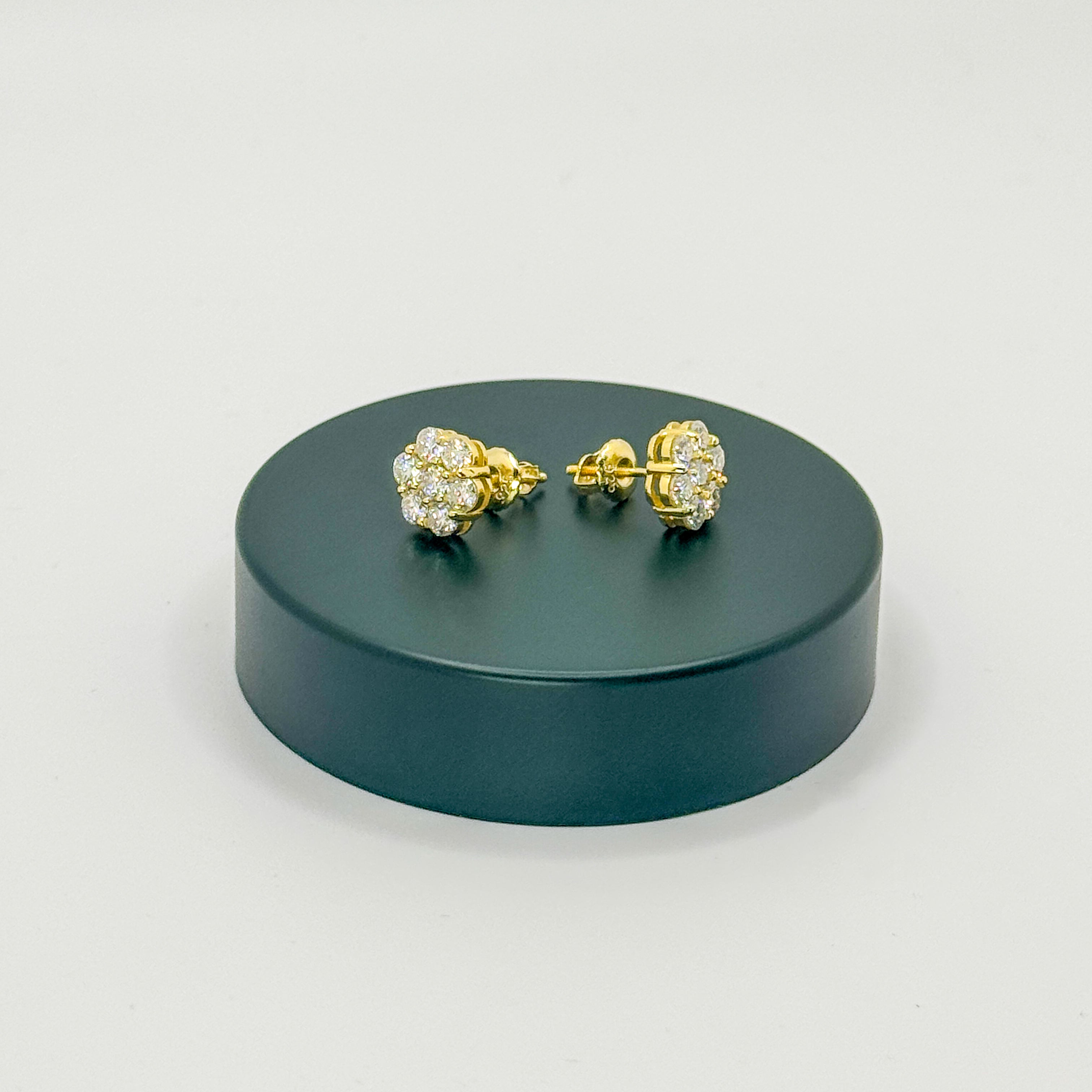 A pair of gold stud earrings with clear pavé gemstones, displayed on a dark green circular stand against a light gray background.