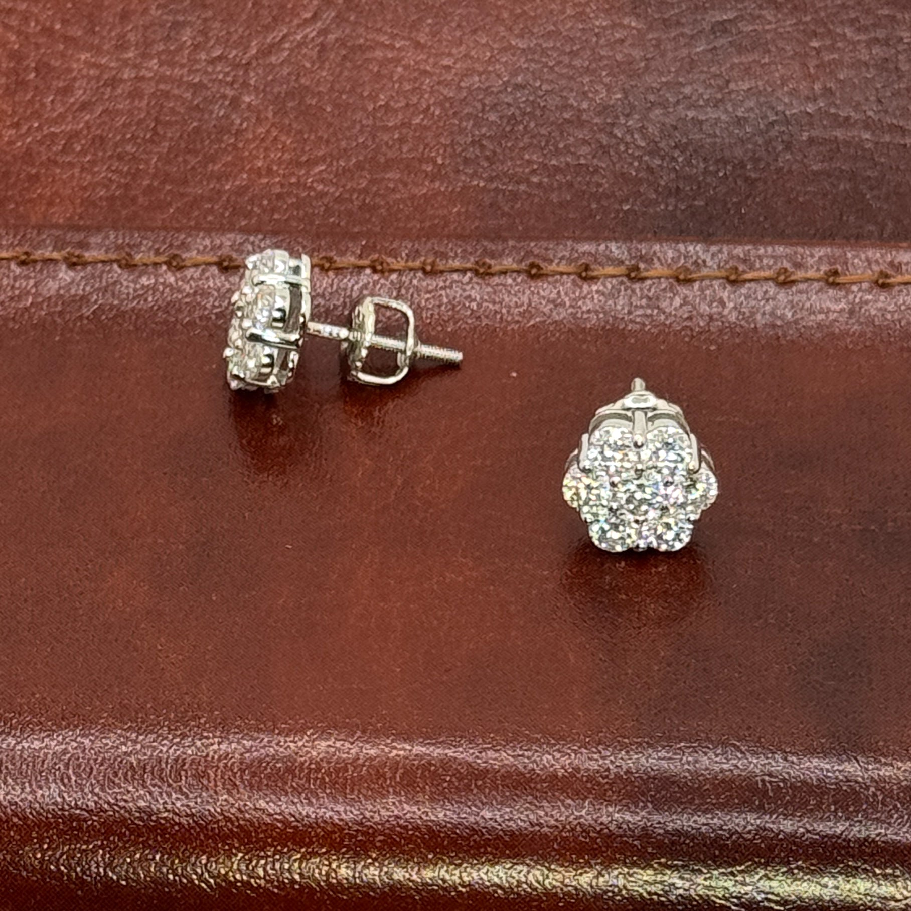 A pair of silver-toned stud earrings with pavé-set clear stones, shown on a brown leather surface. One earring is displayed from the front and the other from the side/back.